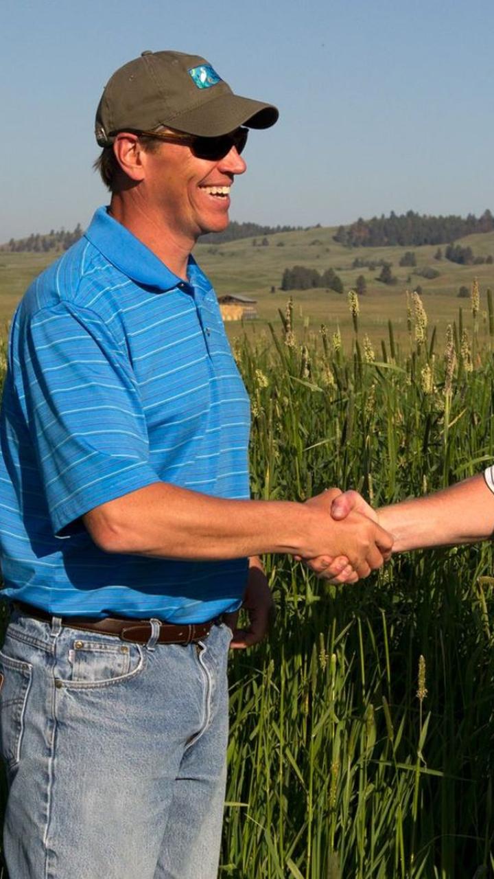 Two smiling men shaking hands in a field of tall grass in Montana's Blackfoot Valley.