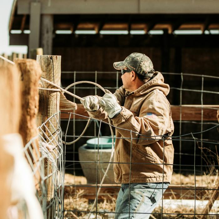 A man in a brown jacket working at a gray metal fence outdoors during daytime.