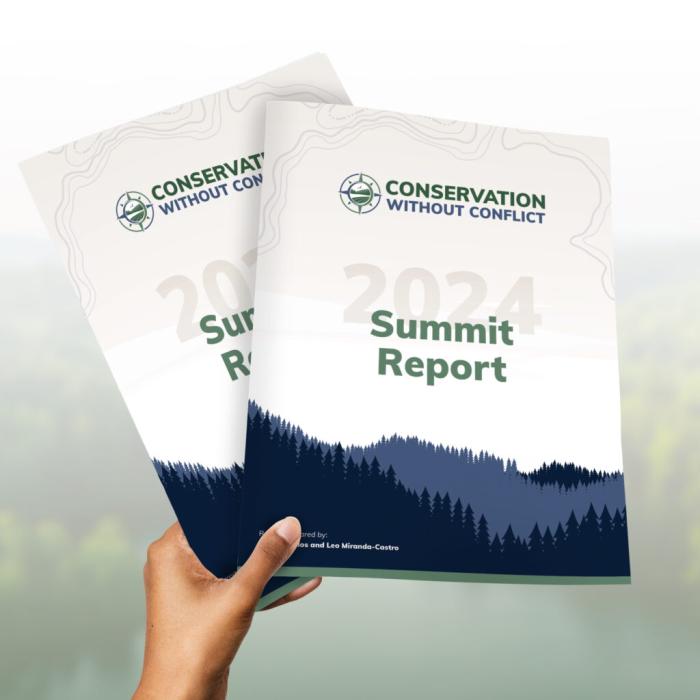 A hand holding two printed copies of the Conservation Without Conflict 2024 Summit Report, with a blurred forest and river landscape in the background.